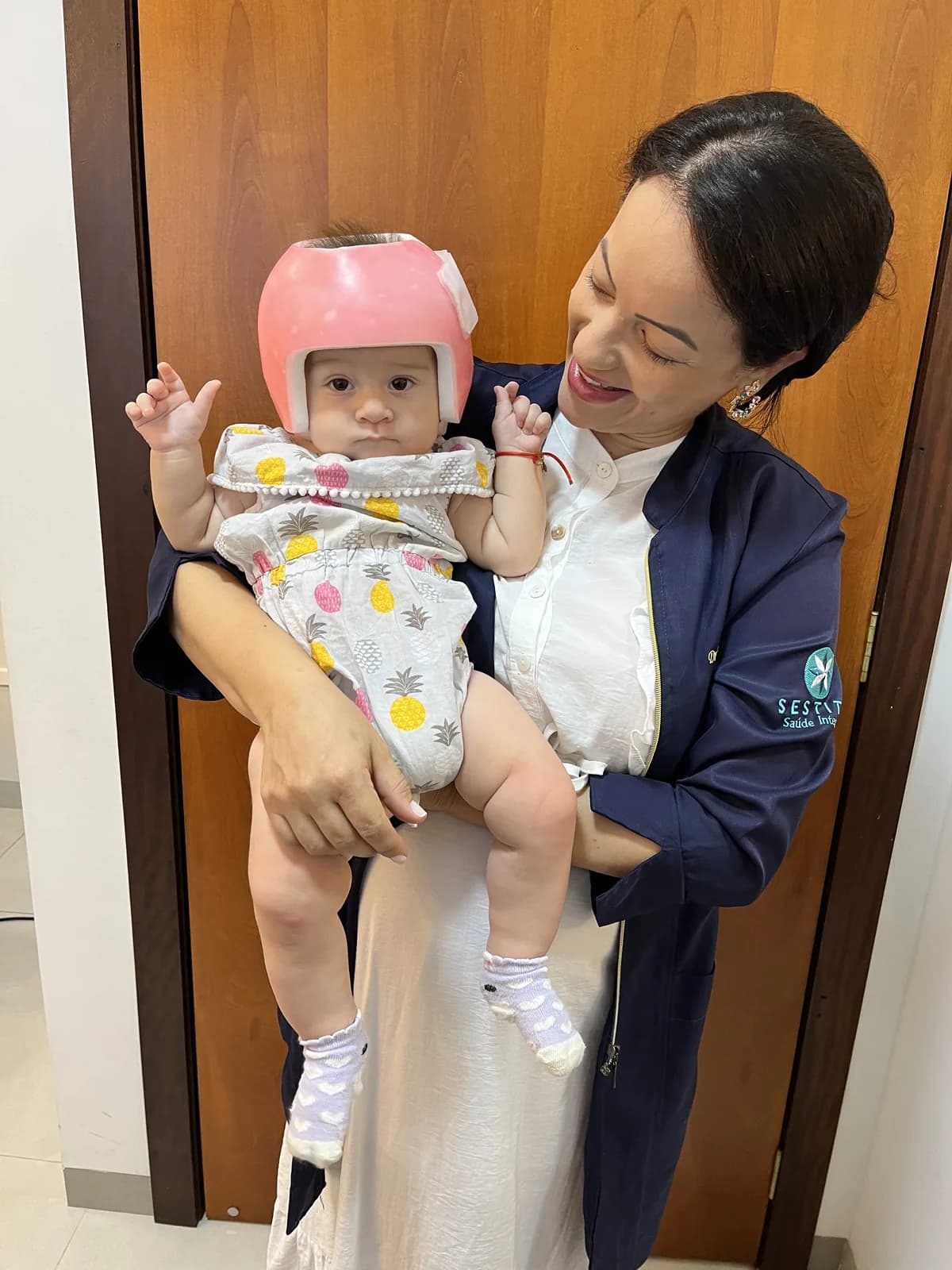 Bebê usando capacete craniano (órtese craniana) cor-de-rosa no colo da Dra. Ligiane Gama